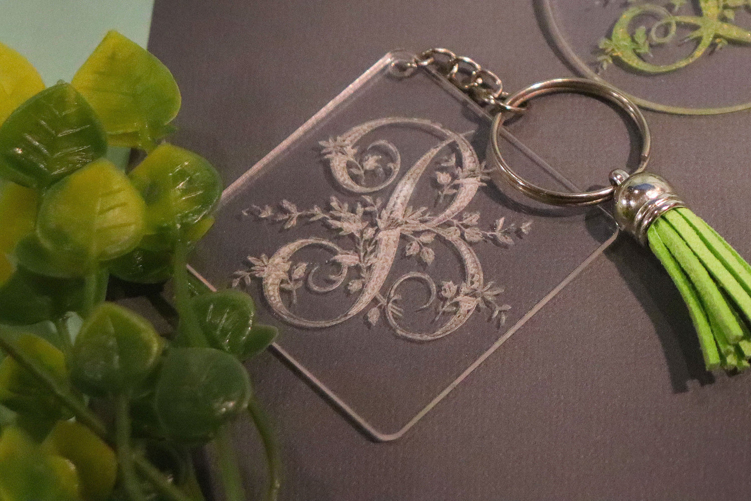 Three engraved acrylic ikeychains with monogram designs and green tassels on a dark surface.